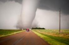 A massive tornado that hit near Dodge City, KS in 2016, at the end of a long dirt road.  Three cars are stopped on the road in front of the twister.  The funnel cloud is wider than the road itself, and you can see the dust and debris whirling around the base.