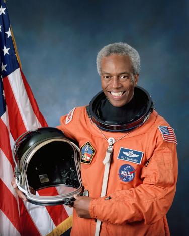 Guion Bluford's official NASA photo. He wears an orange suit and holds his helmet. The American flag is on the left side (to the viewer) of the photo and Bluford is smiling. 