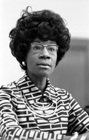Headshot of Shirley Chisholm. She wears glasses, earrings, a stylish hairdo, and a dress with a block design.