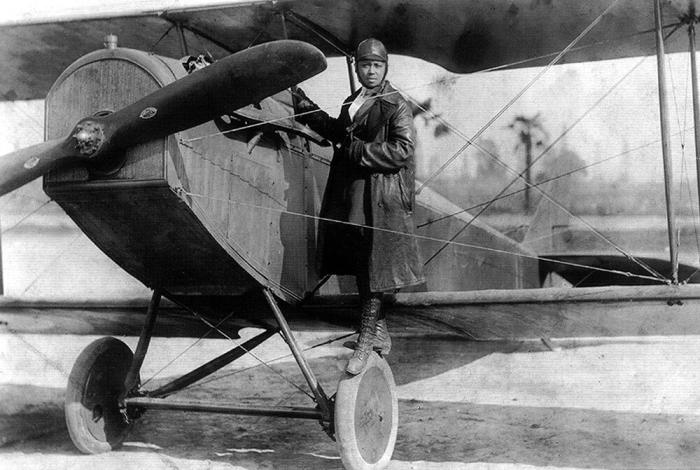 Black and white photo of Bessie Coleman standing on the wheel of her plane.