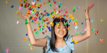 Tween/Teen girl with long black hair and a blue shirt is surrounded by origami paper cranes