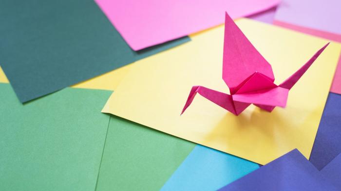A pink origami crane sits atop origami paper in multiple colors